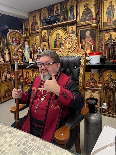 homem de veste vermelha com báculo, sentado numa cadeira dourada, cercado de santos e ícones religiosos — texto: SACRED